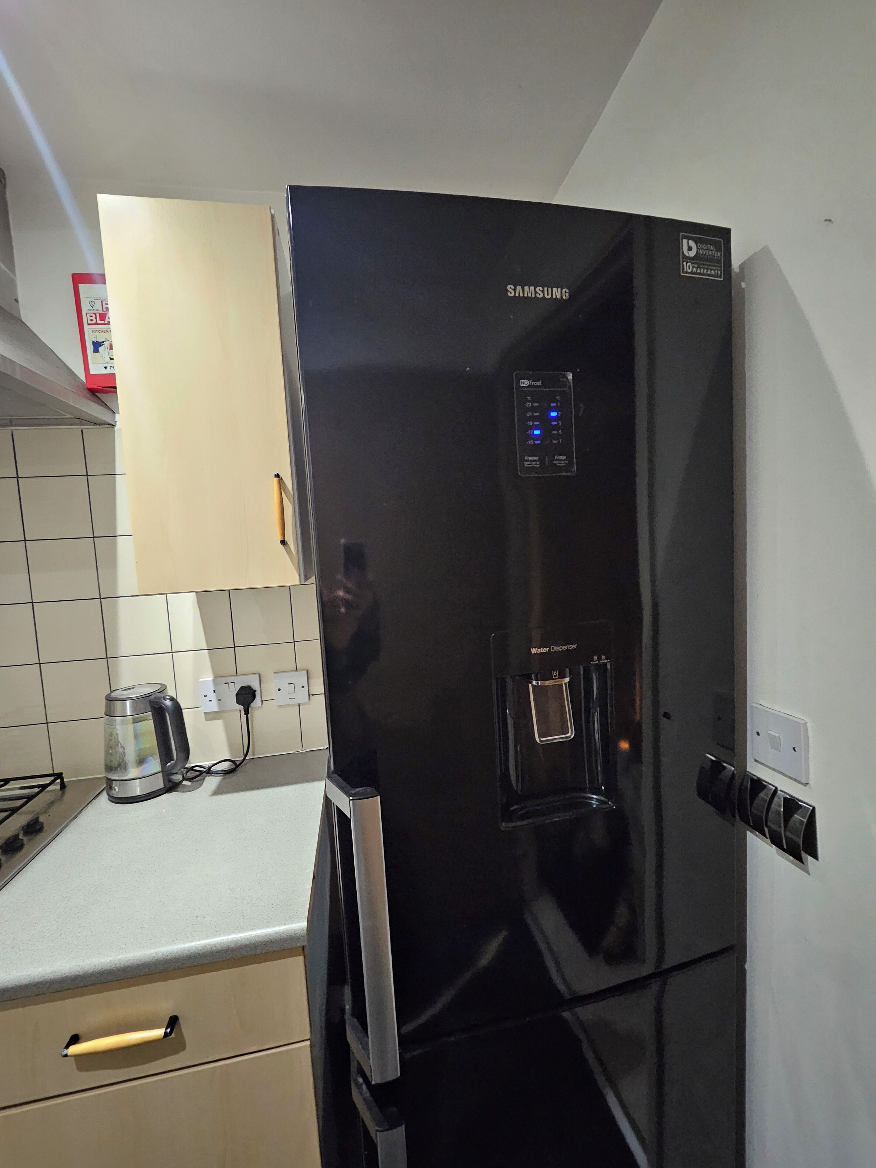 Un refrigerador Samsung negro con dispensador de agua, una tetera y un armario encima.