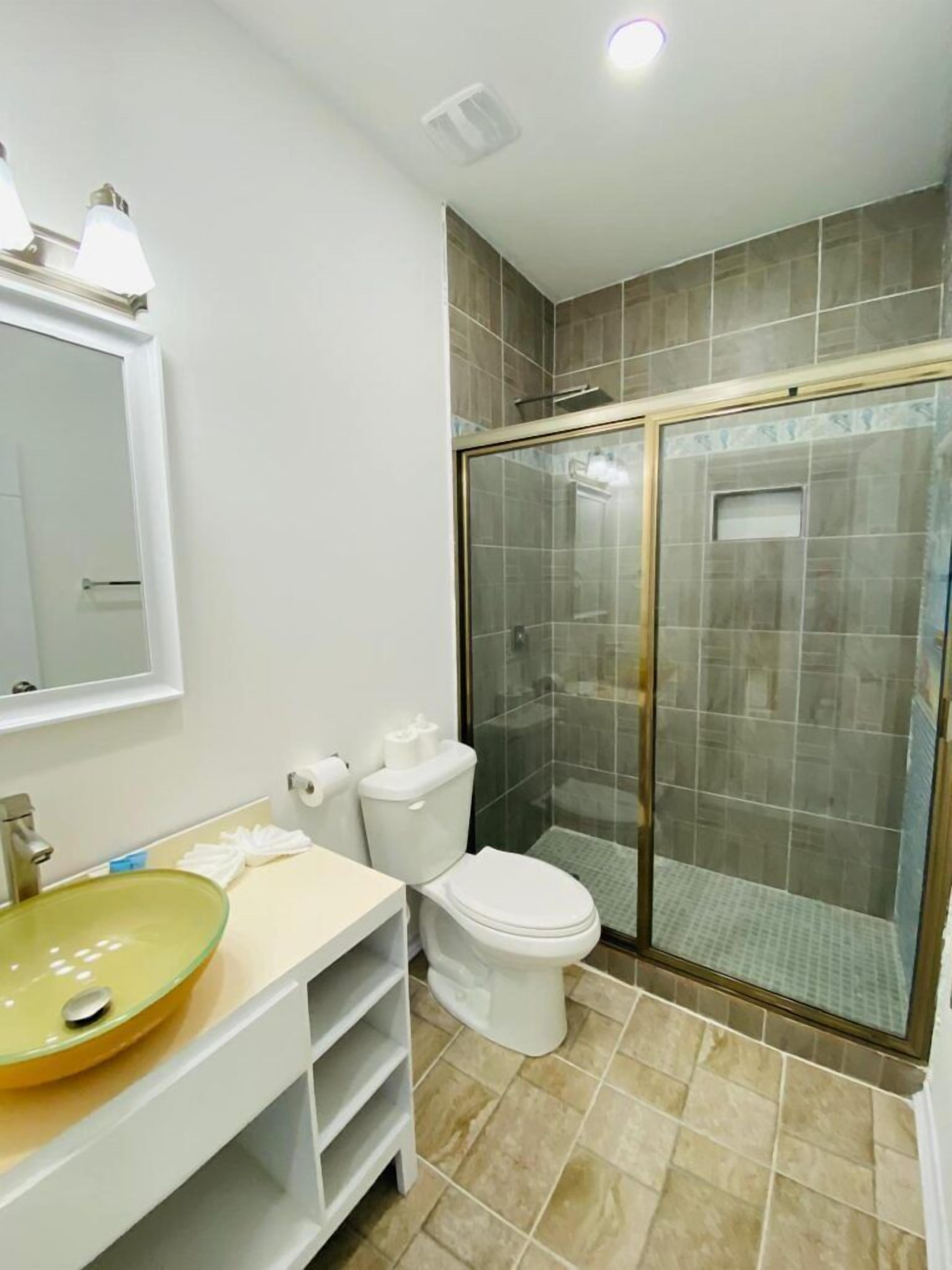 Un baño con ducha envuelta en vidrio, un inodoro blanco, un lavamanos amarillo y un espejo sobre el lavamanos.