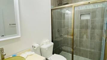 Un baño con ducha envuelta en vidrio, un inodoro blanco, un lavamanos amarillo y un espejo sobre el lavamanos.