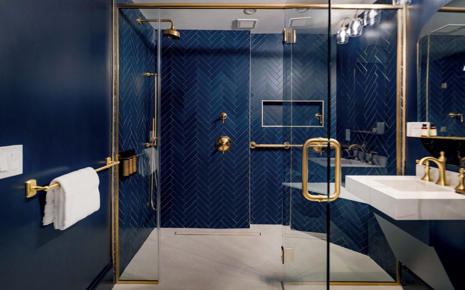 Un baño moderno con ducha envuelta en vidrio, accesorios dorados y pared con estampado geométrico azul oscuro.