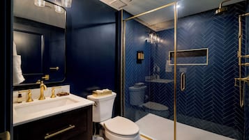 A modern bathroom with a dark blue herringbone tile wall, a glass-enclosed shower, and gold fixtures.