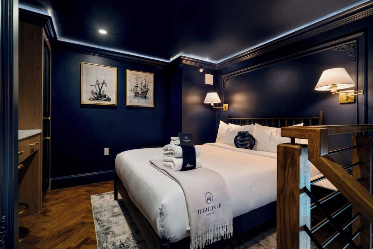 Ein Hotelzimmer mit dunkelblauer Wand, einem Bett mit weißer Bettwäsche, einem Holz-Kopfteil, einem Nachttisch mit Lampe und gerahmten Kunstwerken an der Wand.