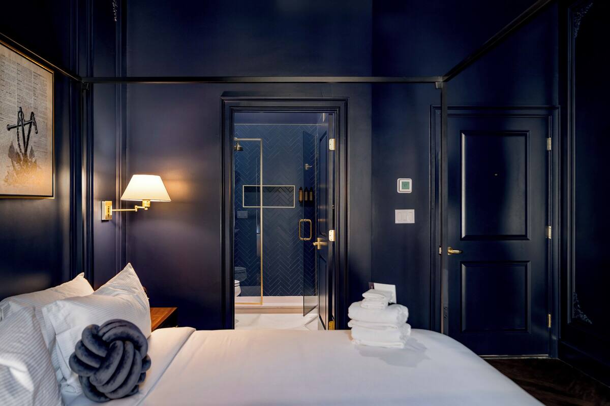 Ein dunkel eingerichtetes Hotelzimmer mit einem Bett, einer Wandlampe, einer Tür mit Glasfenster und einem gerahmten Bild an der Wand.