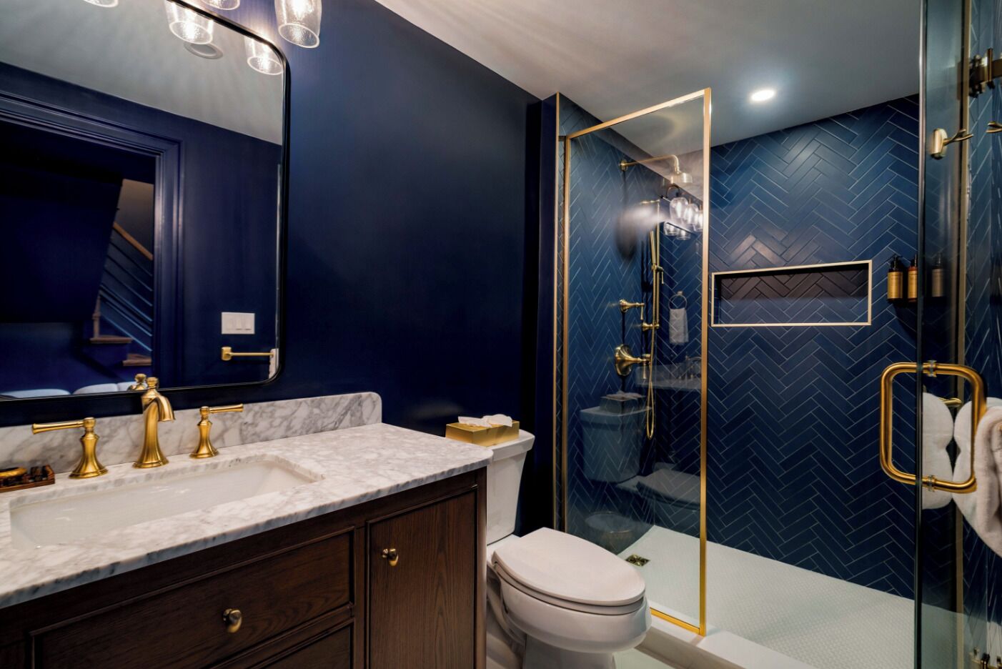 Un baño moderno con pared de azulejos estampados en diseño de espiga color azul oscuro, ducha envuelta en vidrio y tocador de mármol con detalles dorados.