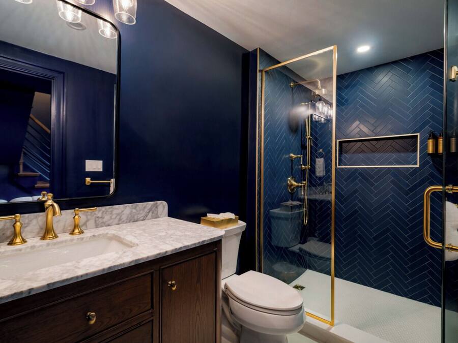 A modern bathroom with a dark blue herringbone tile wall, a glass-enclosed shower, and a marble vanity with gold fixtures.