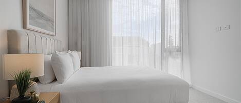 Un dormitorio moderno con una cama grande, una mesita de noche con lámpara, un cuadro enmarcado en la pared y cortinas traslúcidas en la ventana.