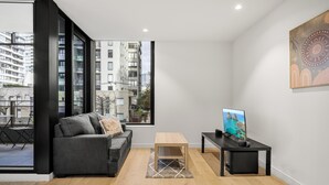 A modern living room with a grey sofa, a wooden coffee table, a black TV stand, and a large window with a city view. - Chic 1-bed South Melbourne Unit With Pool & Gym (Melbourne)