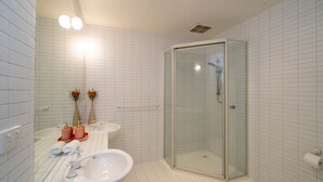 Ein modernes Badezimmer mit weiß gefliesten Wänden, einer in Glas abgeschlossenen Dusche, einem weißen Waschbecken und zwei Vasen mit Blumen.