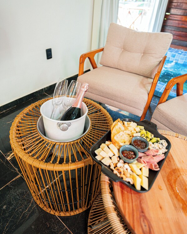A wicker table with a bottle of champagne and glasses, a tray of cheese and meats, and a chair in the background.