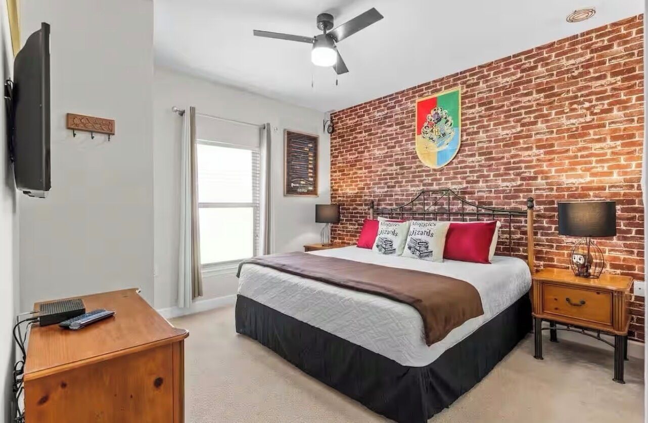 A bedroom with a brick-patterned wall, a bed with decorative pillows, a ceiling fan, a TV, and a bedside table with a lamp.