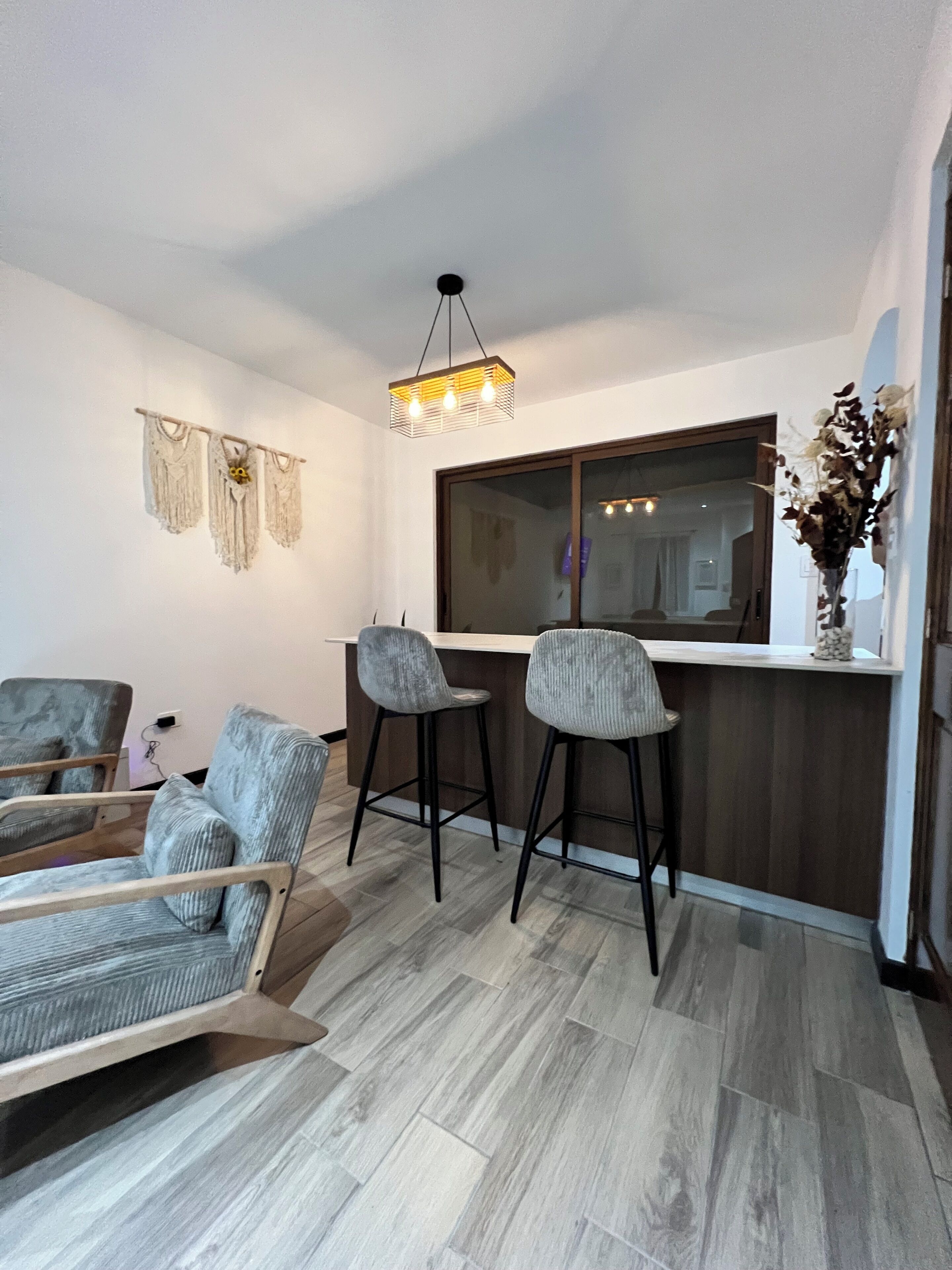 Een moderne woonkamer met houten vloer, een bank, twee barkrukken, een houten salontafel, een hanglamp en een groot raam.