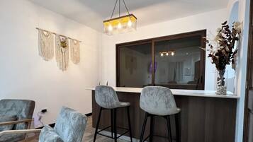 Een moderne woonkamer met houten vloer, een bank, twee barkrukken, een houten salontafel, een hanglamp en een groot raam.