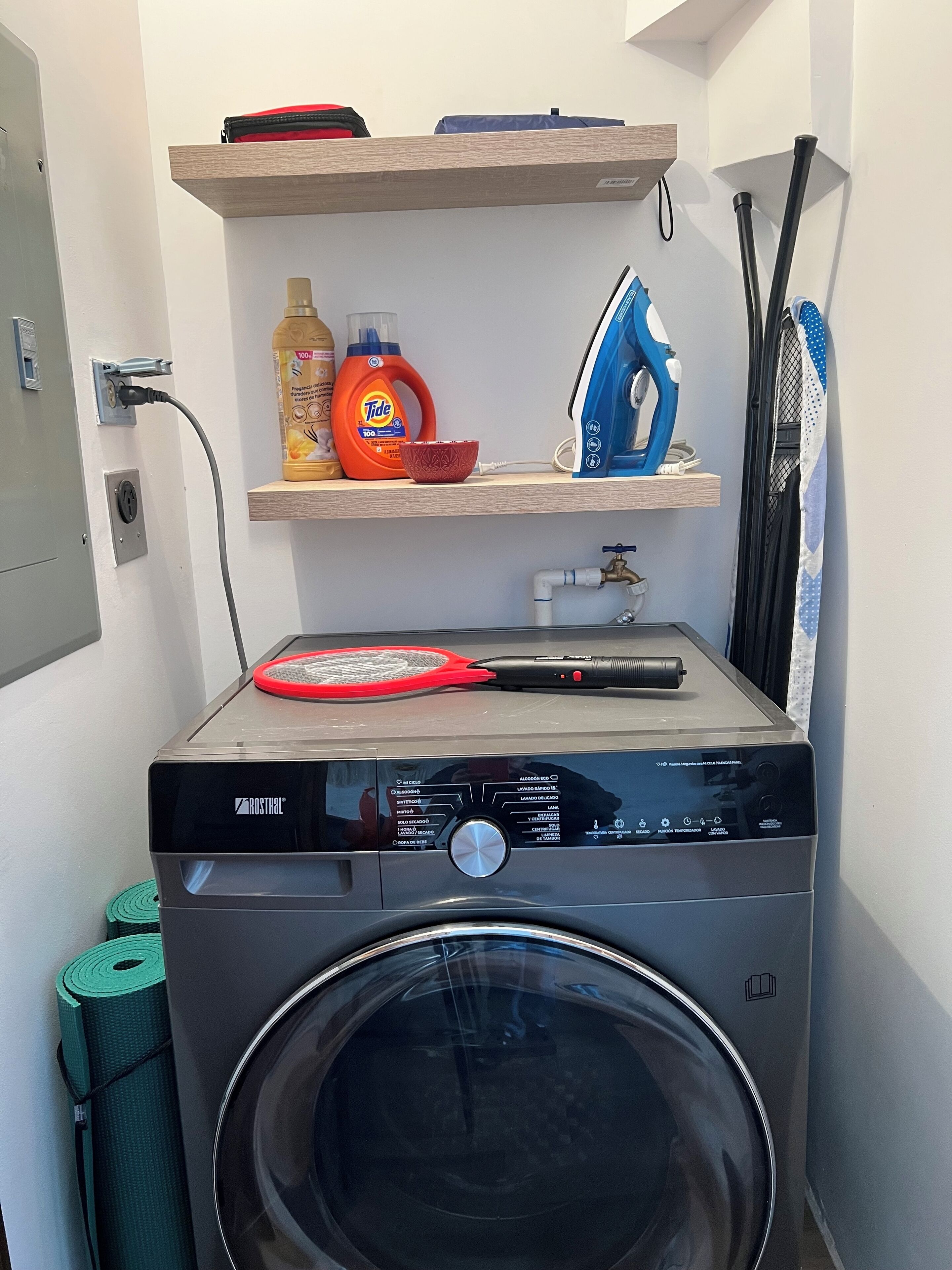 A compact laundry area with a front-loading washing machine, a shelf with cleaning supplies, and a hanging rack.