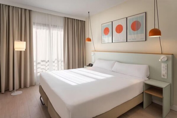 Ein Hotelzimmer mit einem großen Bett, einem Nachttisch, einer Lampe und einem Fenster mit Vorhängen.