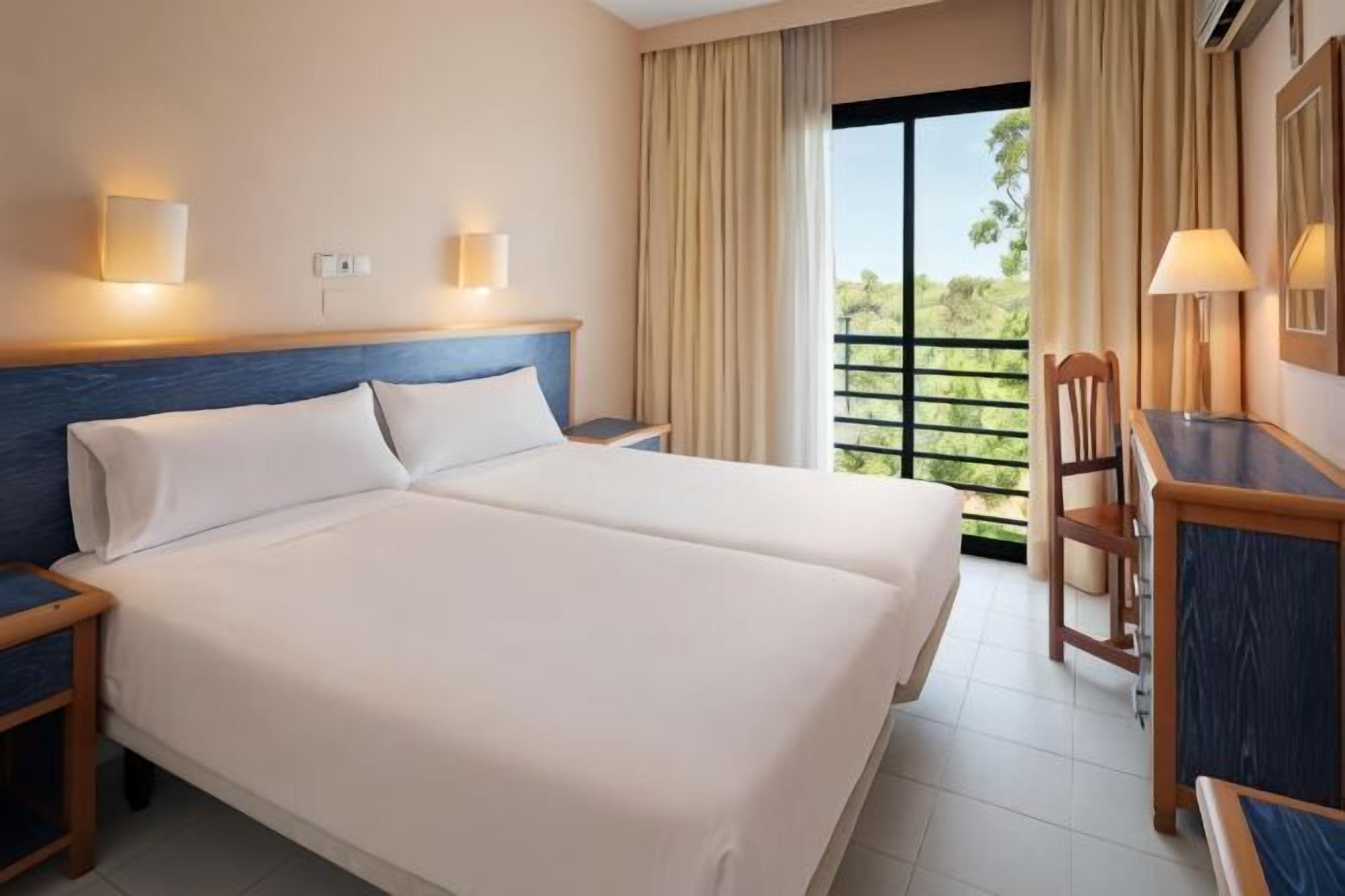 A double bed with two pillows, a nightstand, a desk with a chair, and a window with curtains.