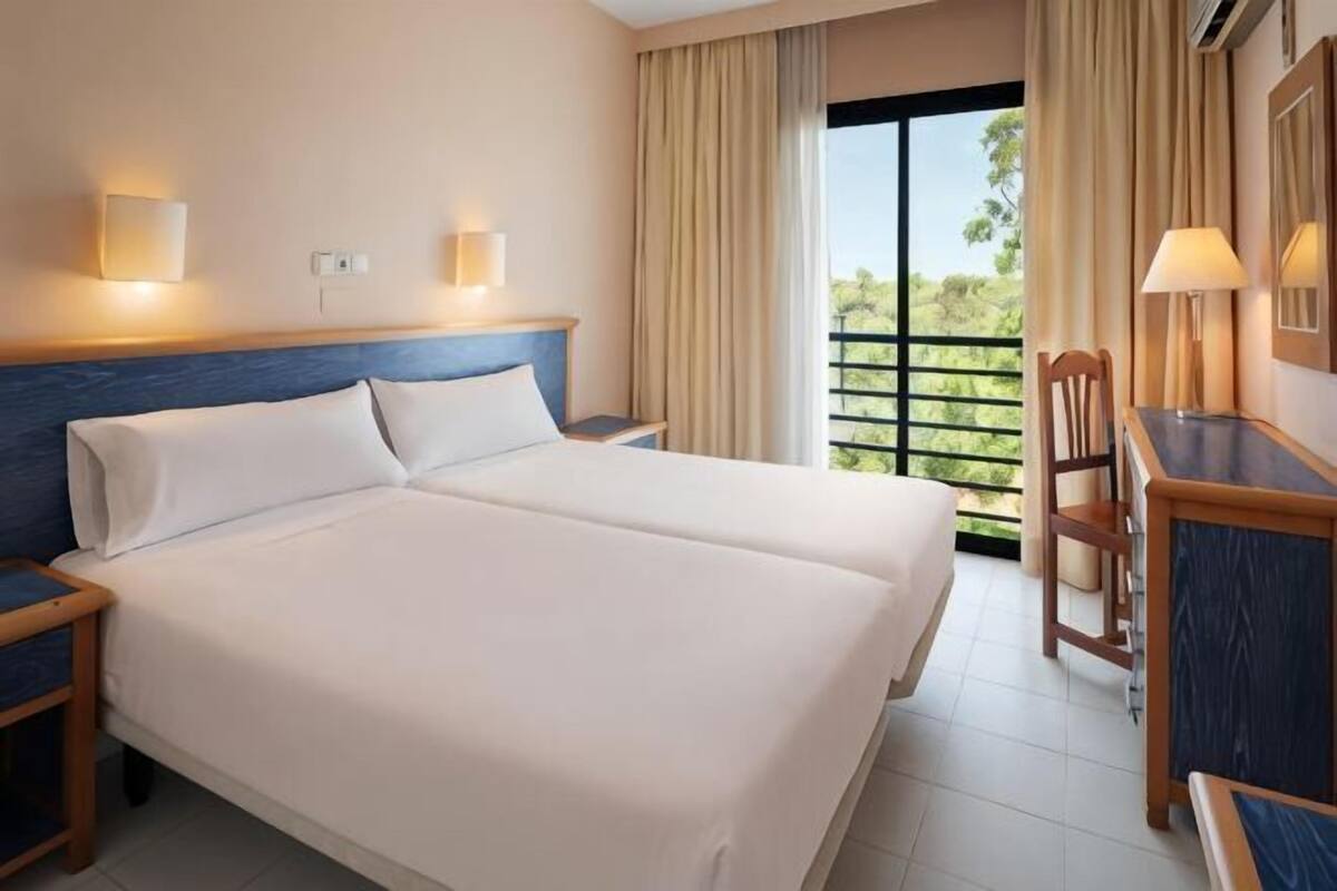 A double bed with two pillows, a nightstand, a desk with a chair, and a window with curtains.