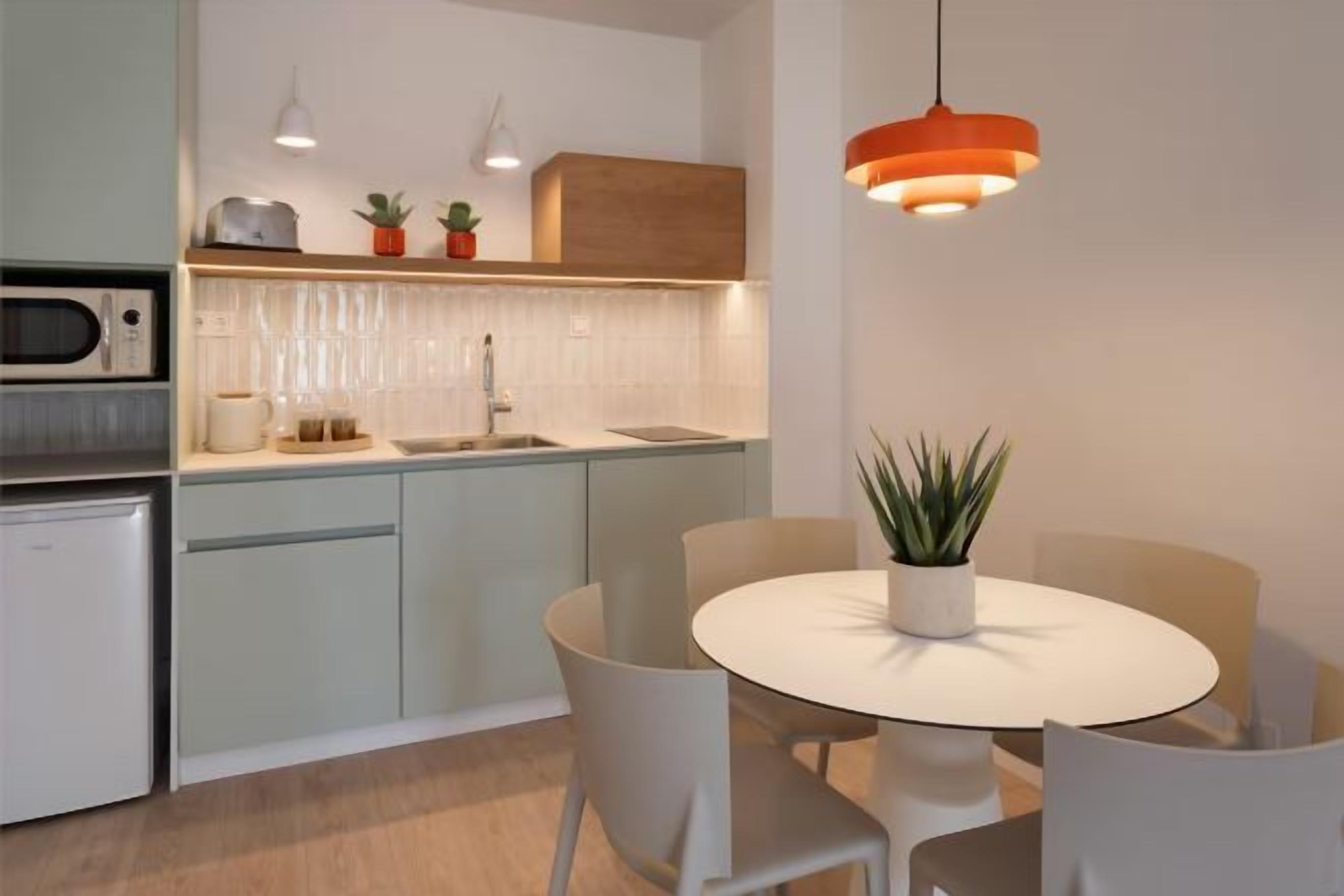 A modern kitchen with a built-in microwave, a sink, and a countertop with a toaster and potted plants. There is a round dining table with chairs and a hanging orange pendant light.