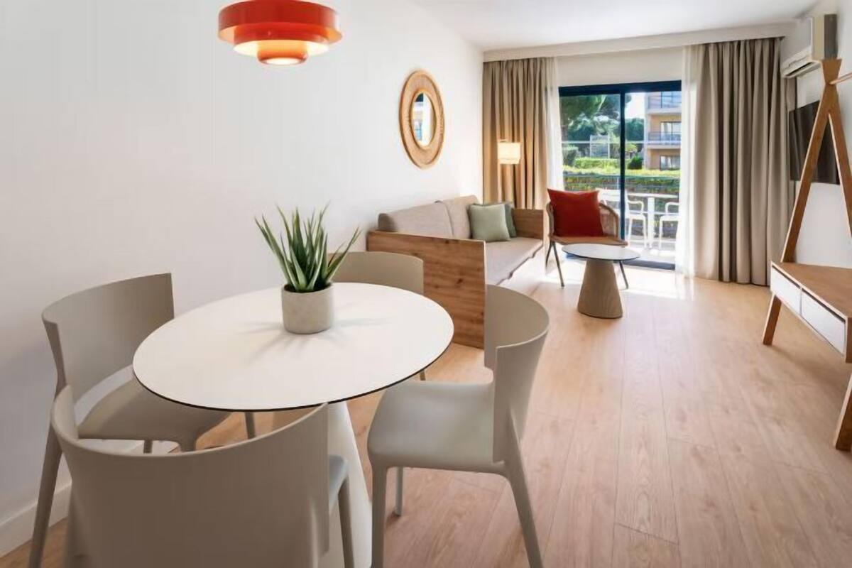 A modern hotel room with a round dining table, two chairs, a sofa, and a small wooden side table.
