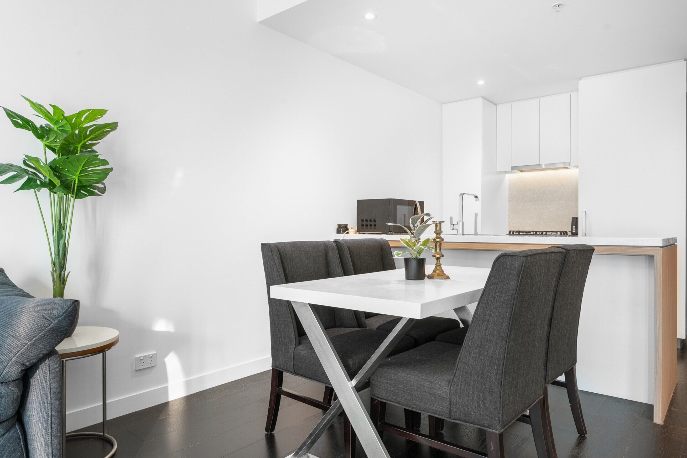 A modern dining area with a white table and grey upholstered chairs, a potted plant, and a kitchen in the background.