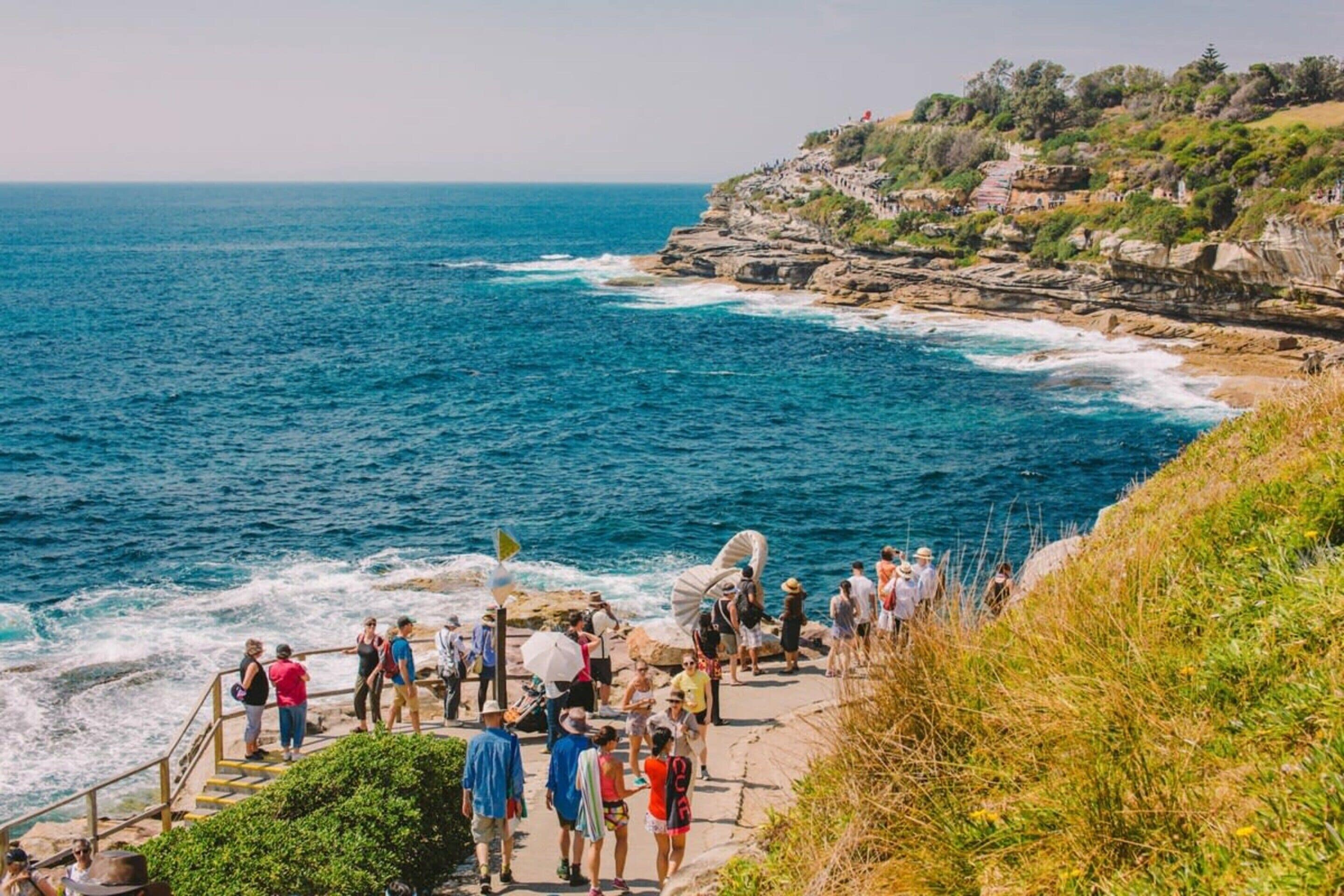 A coastal walkway with people walking, overlooking the sea and rocky cliffs.