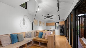 Uma sala de estar moderna com piso de madeira, um sofá embutido com almofadas, um ventilador de teto e uma cozinha visível por uma porta aberta.