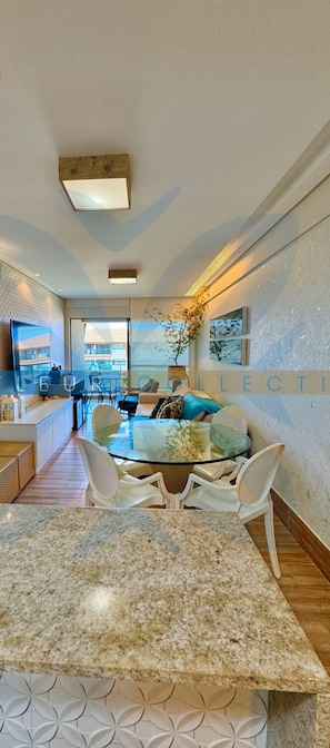 Um interior moderno com uma sala de jantar com mesa de vidro e cadeiras brancas, uma cozinha com forno embutido e uma sala de estar com sofá e mesa de centro.