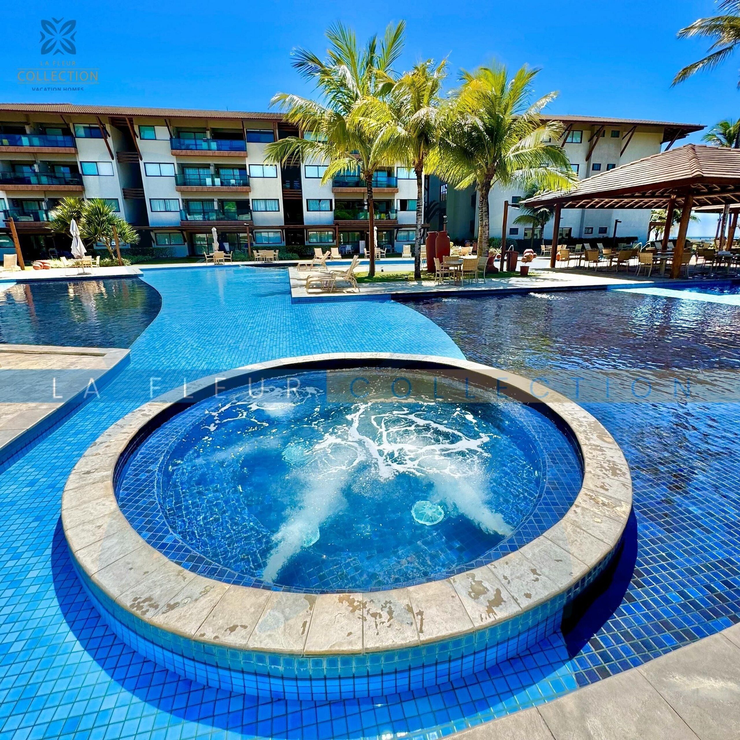 Una piscina con un jacuzzi en el centro, rodeada de sillones de descanso y palmeras.