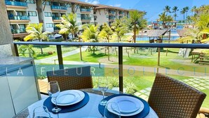 A dining table set with plates and wine glasses on a balcony overlooking a resort area with palm trees and a pool. - Luxury Pool View at La Fleur Polynesia (Porto de Galinhas)