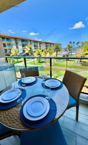Een eettafel gedekt met borden en wijnglazen op een balkon met uitzicht op een resort, met palmbomen en een zwembad.