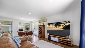 A modern living room with a leather sofa, a dining area with chairs, a TV on a wooden stand, and a large window with a view. - Family-friendly Home With Private Pool and BBQ (Sunshine Coast)