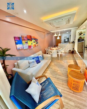 A modern living room with a sofa, a dining table, and chairs, a kitchen area, and a balcony with orange wicker stools.