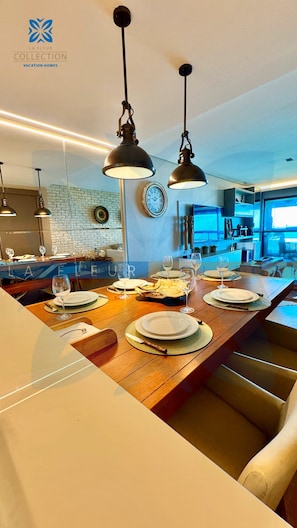 A modern dining area with a wooden table set for a meal, pendant lights, and a kitchen in the background.