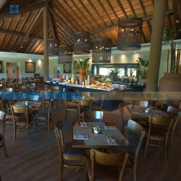Un restaurant avec des tables et des chaises en bois, un espace buffet proposant divers plats, et des lampes en osier suspendues au plafond.