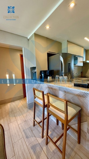 A modern kitchen with a built-in stove, coffee maker, and a sink. There are two wooden chairs with woven seats. The floor is tiled, and the walls are painted in a light color.