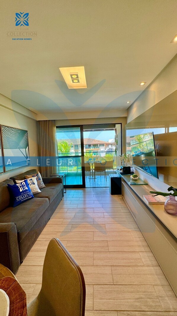 A modern living room with a sofa, a coffee table, and a TV. There is a balcony with a view of buildings and greenery.