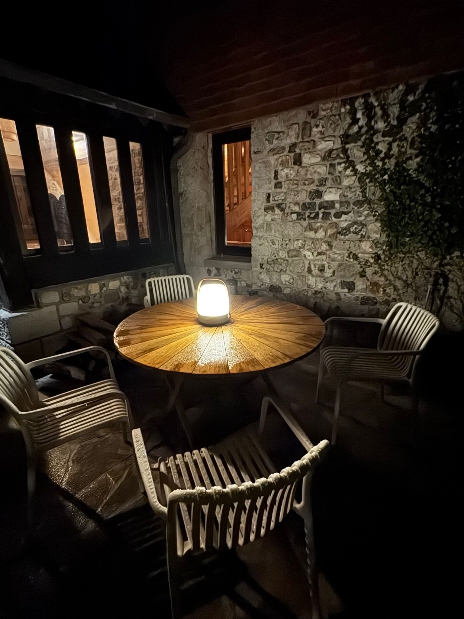 Une table ronde en bois, surmontée d’un luminaire central, est entourée de six chaises en osier. L’ensemble est agrémenté d’un mur en pierre et d’une fenêtre avec des stores verticaux.