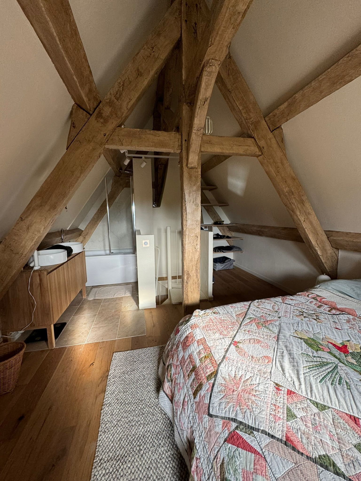 Une chambre avec des poutres apparentes, un lit recouvert d’une courtepointe à motifs, une chaise blanche et une petite armoire en bois.