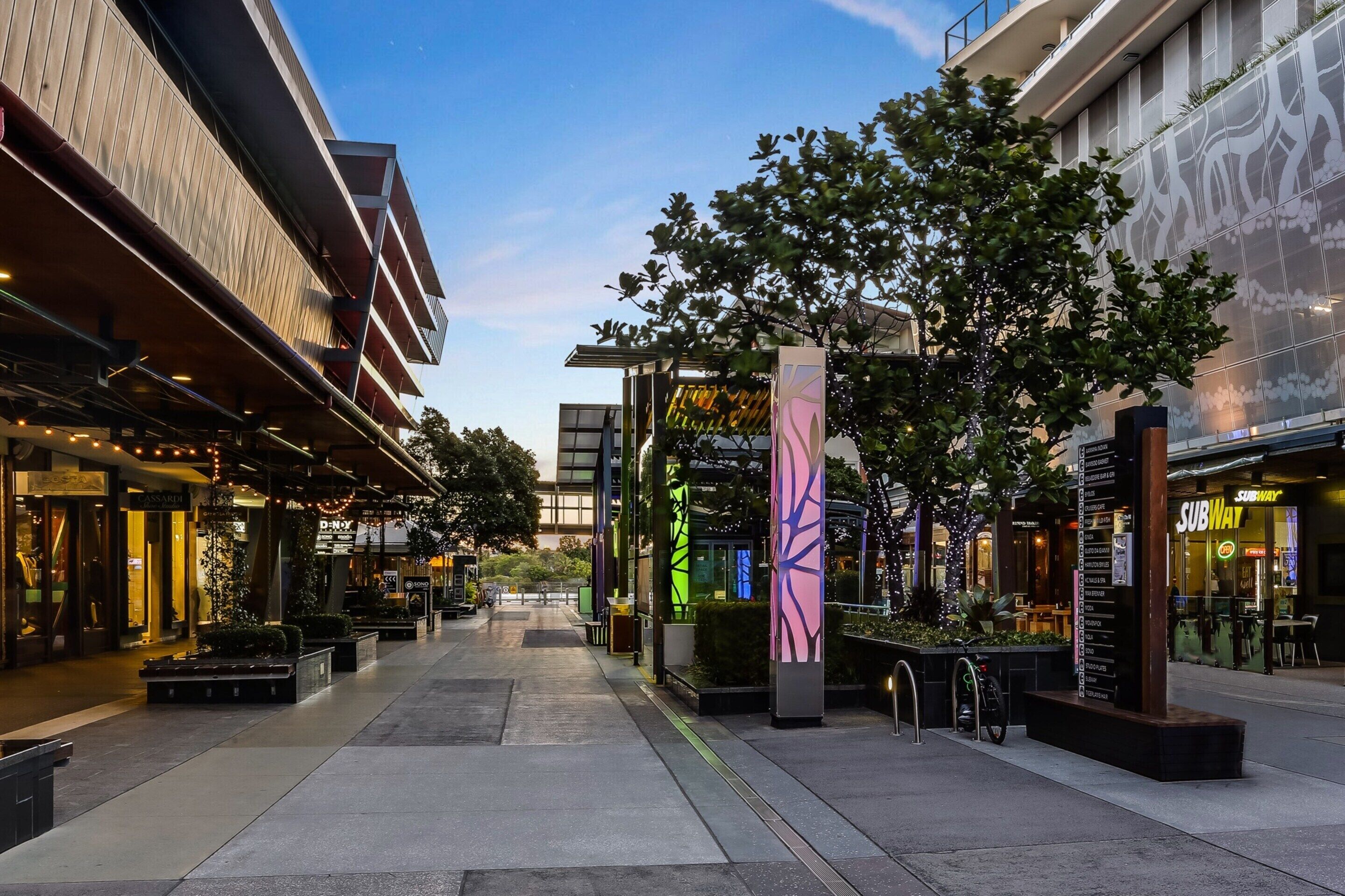 A modern urban street with shops, a pedestrian walkway, and a tree-lined path.