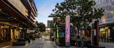 A modern urban street with shops, a pedestrian walkway, and a tree-lined path.