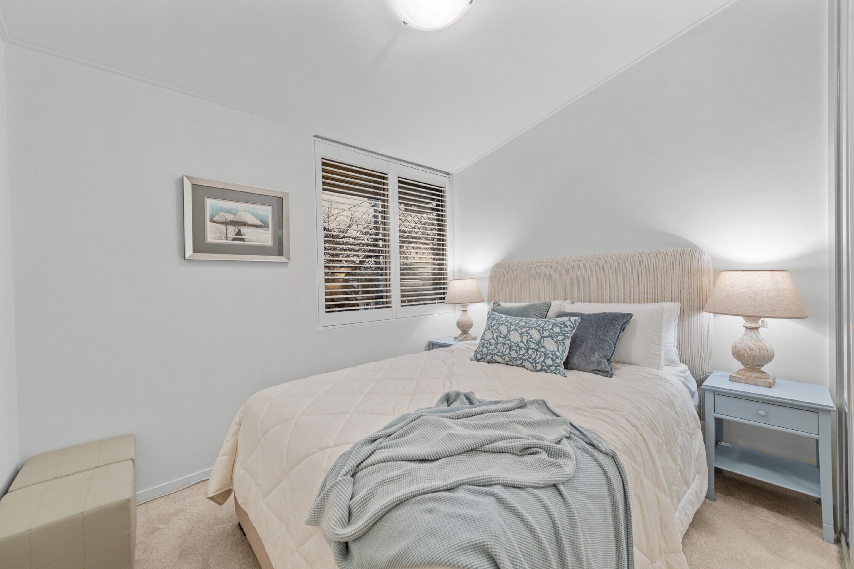 A bedroom with a large bed, bedside lamps, a window with blinds, a framed picture on the wall, and a nightstand.