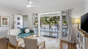 Un salon avec un canapé vert, deux fauteuils, un téléviseur et une grande fenêtre donnant sur la rue.
