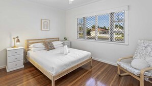 Une chambre à coucher avec un lit en bois, des tables de chevet blanches, une lampe de bureau, une chaise à motifs végétaux, une fenêtre avec vue et un ventilateur de plafond.