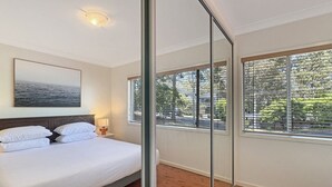 A bedroom with a bed, a nightstand, a lamp, a painting, and large windows with blinds. - Pet-friendly 2-bed Beach House With Garden & BBQ (Surf Beach)