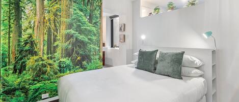 A bedroom with a large forest mural on one wall, a bed with white bedding, and a nightstand with a lamp.