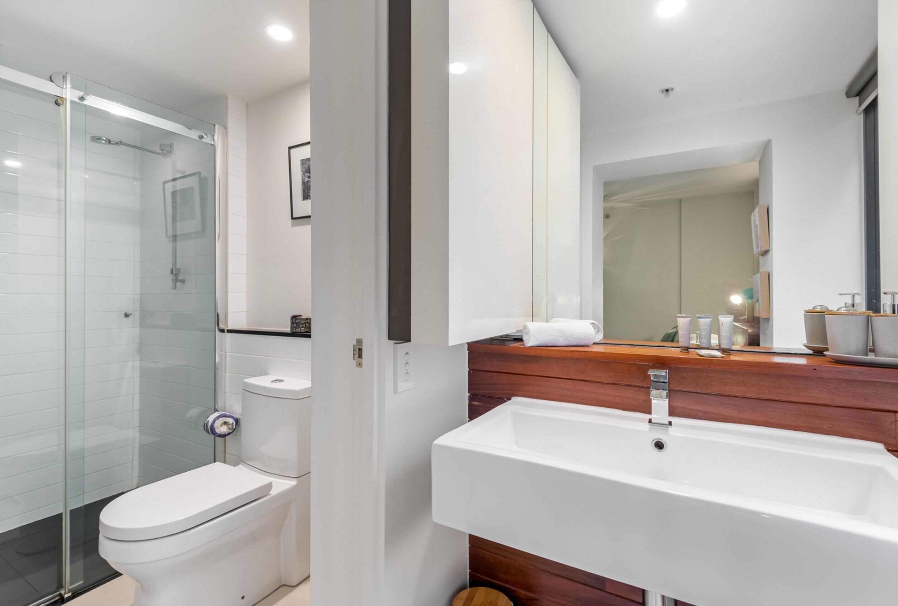 A modern bathroom with a glass shower enclosure, a white toilet, a wooden vanity with a sink, and a large mirror.