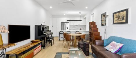 A modern living room with a flat-screen TV, a piano, a dining area, and a staircase leading to an upper level.