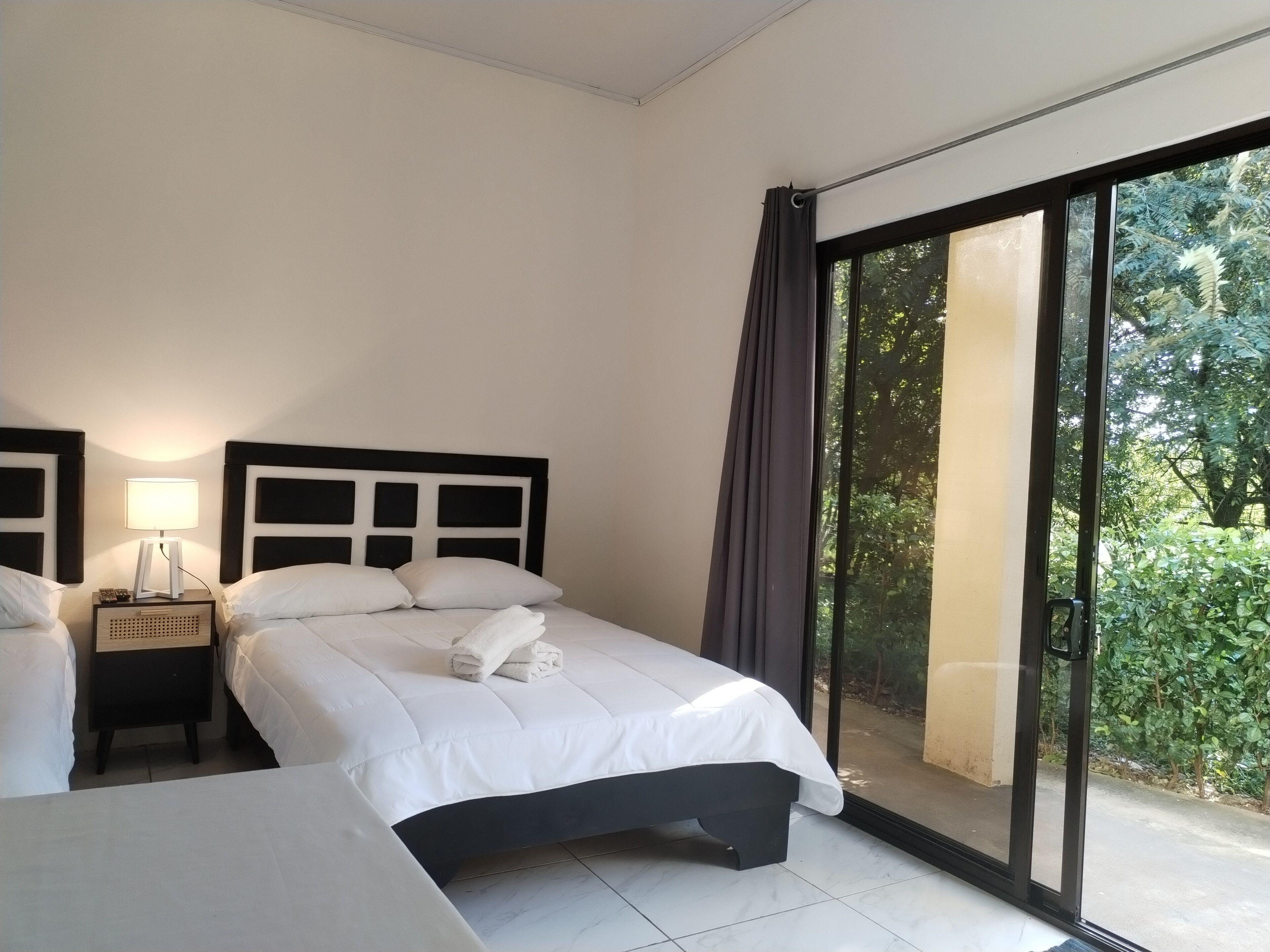 Une chambre d’hôtel avec un grand lit, une table de chevet avec une lampe et une vue sur la verdure grâce à une porte-fenêtre coulissante.