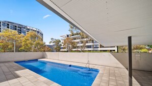 An outdoor swimming pool with a tiled deck, surrounded by buildings and trees. - Elegant 2-bed With Pool, Alfresco Dining & Balcony (Canberra)