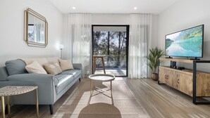 A modern living room with a grey sofa, a round coffee table, a TV on a wooden stand, and a view of a balcony with a plant. - Stylish 1-bed Apartment With Rooftop Access (Perth)