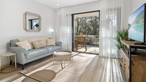 A modern living room with a sofa, a round coffee table, a TV, and a balcony with a view of trees and a lake. - Stylish 1-bed Apartment With Rooftop Access (Perth)